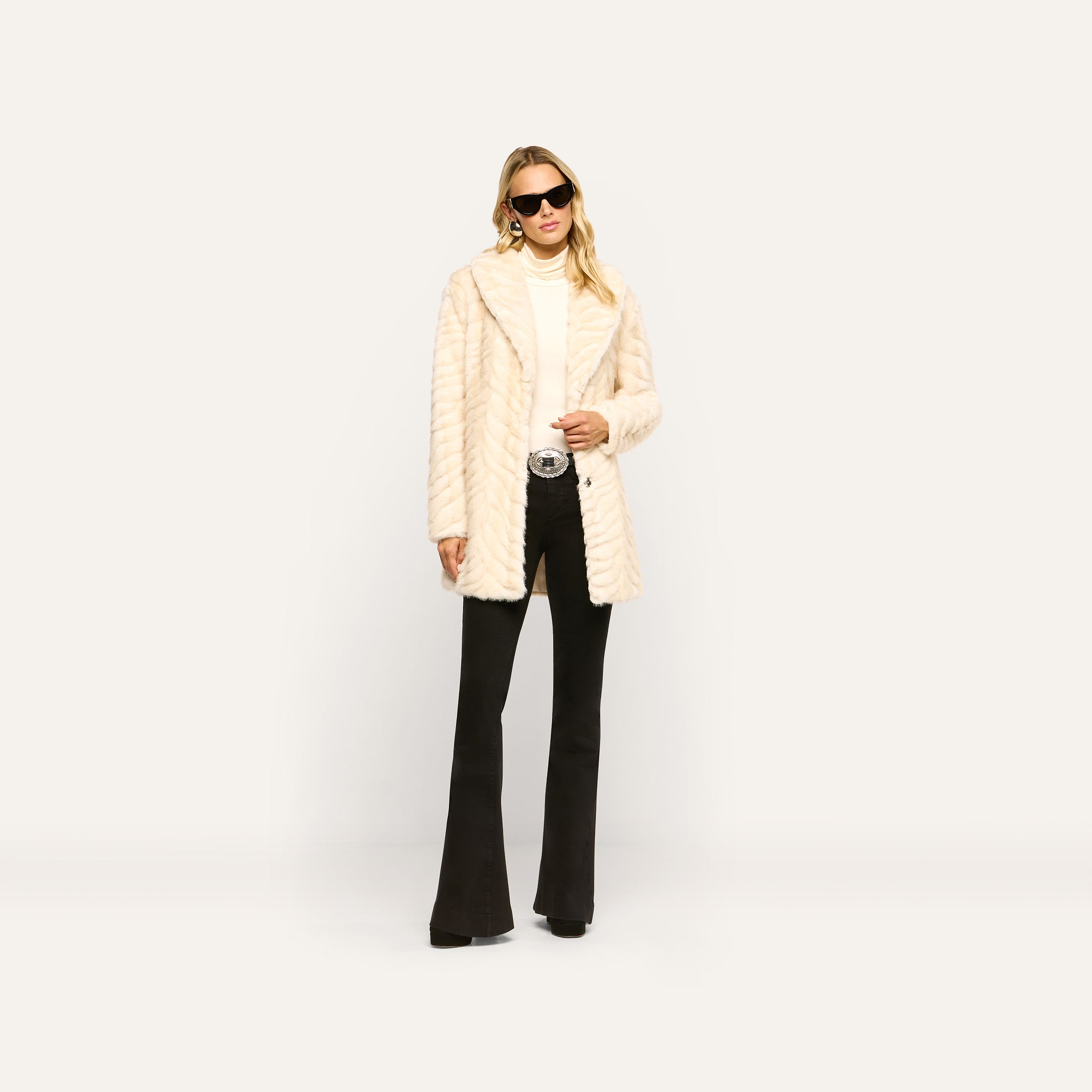 A woman wears a vintage Toira Faux Fur Coat with a chevron pattern over a white turtleneck and black flared jeans, finished with black sunglasses and a large statement belt buckle.