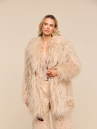 A woman wearing the Toira Faux Fur Coat, a vintage-inspired beige faux fur piece, and sequined pants stands against a plain beige background, gazing slightly to the side with a neutral expression.