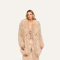 A woman stands facing forward, wearing the Toira Faux Fur Coat—a long, light beige piece—layered over a beige buttoned top and shiny beige sequin pants, channeling a vintage-inspired 70s rockstar vibe against a plain light background.