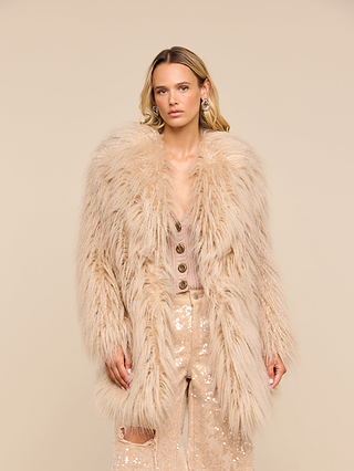 Wearing the Toira Faux Fur Coat, a person with long blonde hair channels a 70s rockstar vibe over a buttoned sequin outfit, posing against a plain beige background.