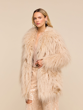 Wearing the Toira Faux Fur Coat, a woman stands against a plain beige background in sequin pants and earrings, channeling a vintage-inspired 70s rockstar vibe as she looks at the camera with a neutral expression.