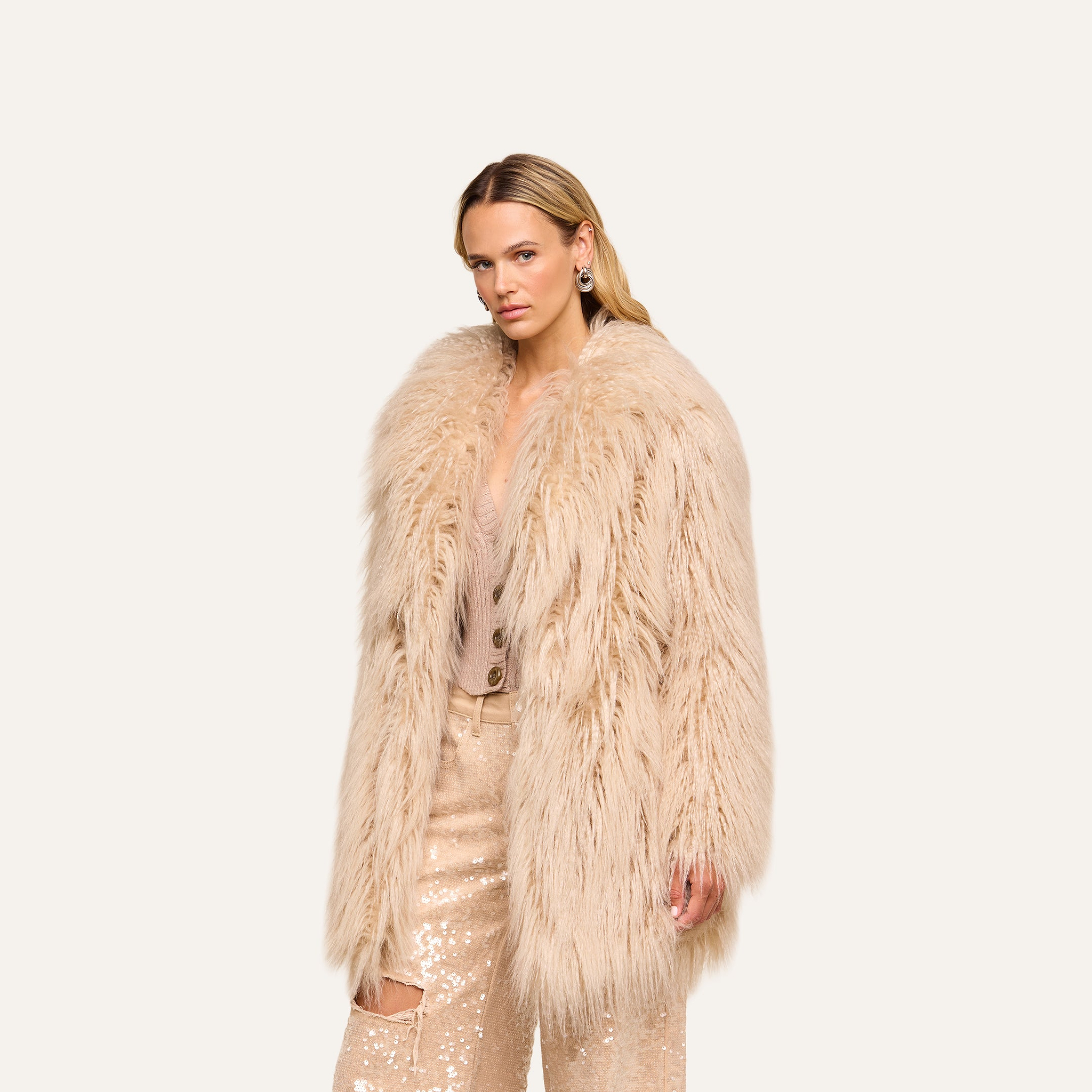 A woman confidently looks at the camera, standing against a plain light background in the Toira Faux Fur Coat—a long, vintage-inspired piece in light beige—layered over a beige sequin outfit.