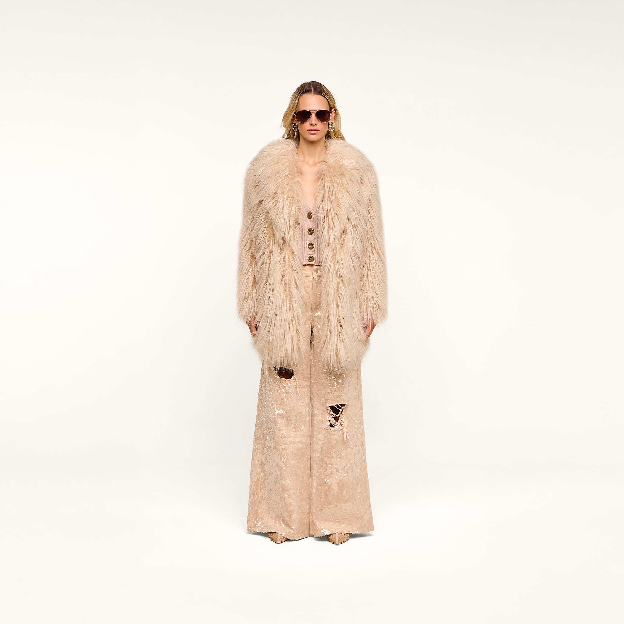 Wearing the Toira Faux Fur Coat, vintage-inspired sunglasses, and matching wide-leg pants with distressed details, a person stands against a plain white background, channeling a 70s rockstar vibe.
