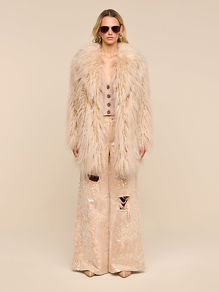 A woman in sunglasses exudes ’70s rockstar vibes as she stands against a plain beige background, wearing the Toira Faux Fur Coat over a matching sequin outfit featuring wide-leg pants with visible rips.