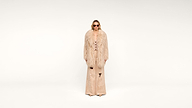 A person wearing sunglasses, a long, beige fur coat, and matching wide-leg pants stands against a plain white background.