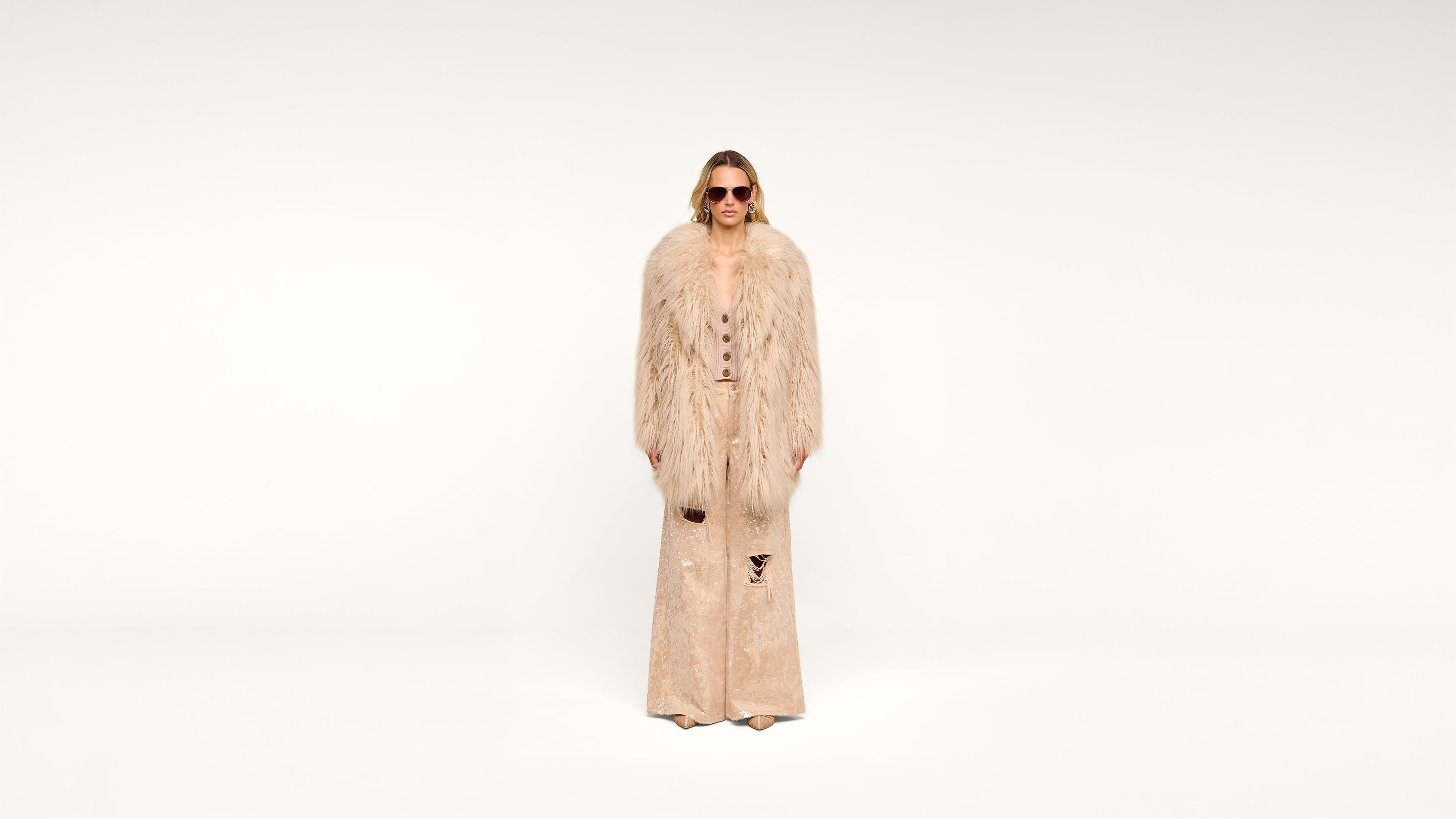 A person wearing sunglasses, a long, beige fur coat, and matching wide-leg pants stands against a plain white background.