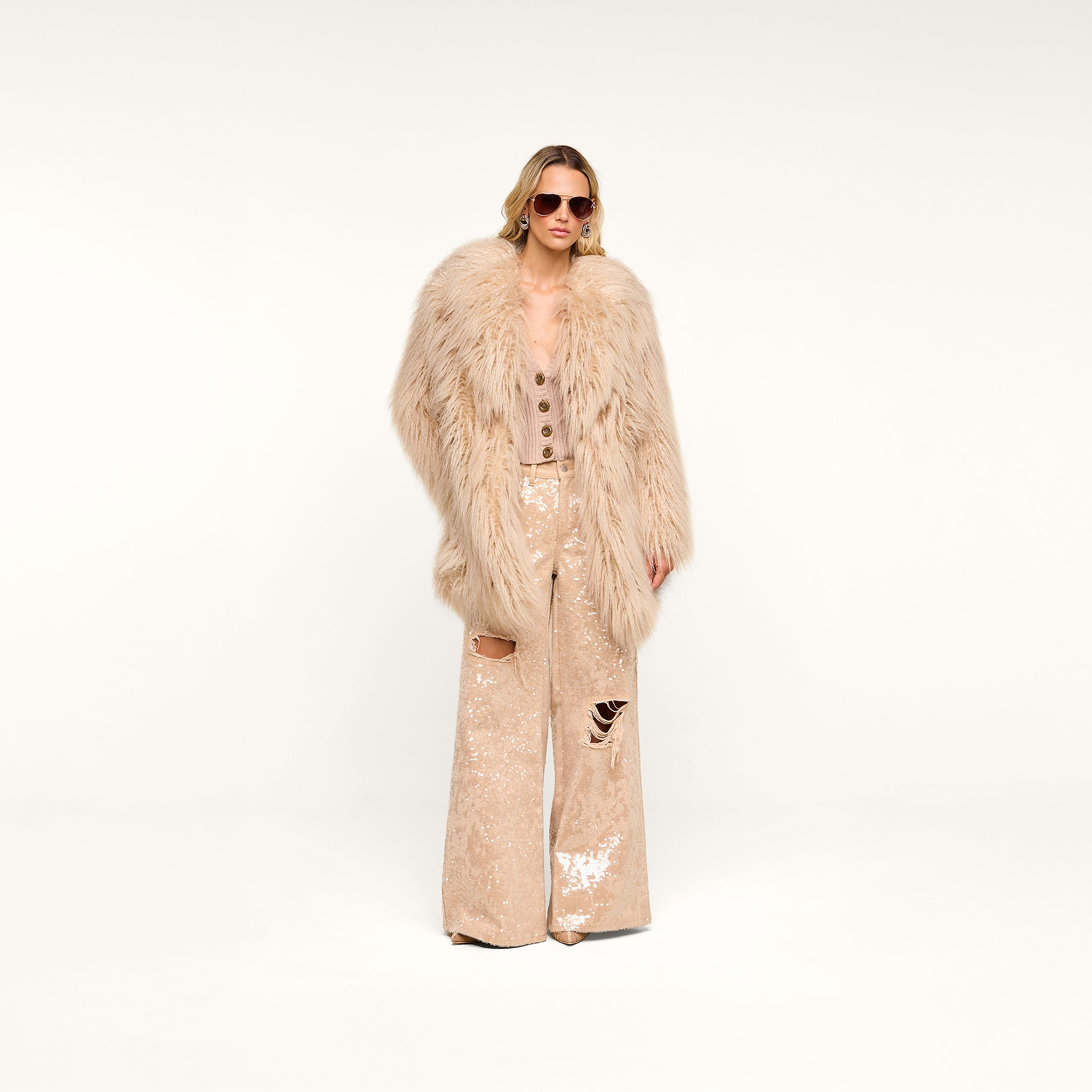 A woman wearing sunglasses, the Toira Faux Fur Coat—a vintage-inspired beige faux fur coat—a beige buttoned top, and beige sequin wide-leg pants with rips stands against a plain white background.