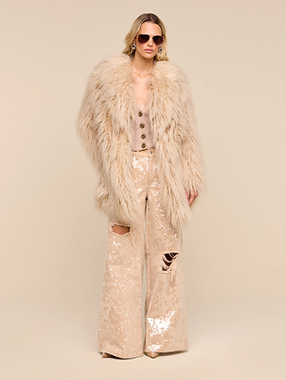 Channeling a 70s rockstar vibe against a beige background, this person wears sunglasses, a vintage-inspired buttoned top, shiny sequined wide-leg pants with rips, and the Toira Faux Fur Coat.