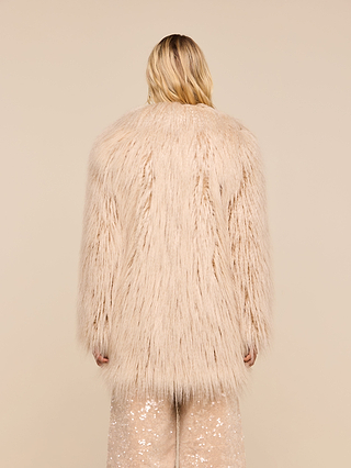 A person with long blond hair is channeling a ’70s rockstar vibe, wearing the Toira Faux Fur Coat in shaggy light beige and sequined pants, standing with their back to the camera against a beige background.