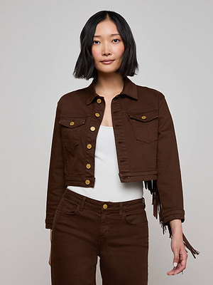 A woman with a medium bob haircut wears the Koda Fringe Jean Jacket, featuring gold buttons, over a white top and matching brown pants, standing against a plain light gray background.