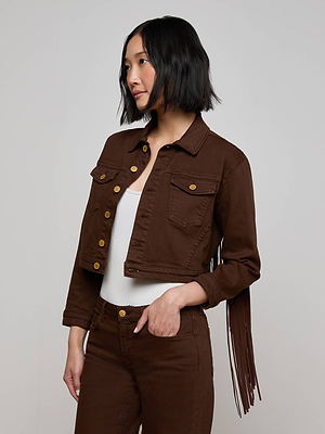 A woman with a bob haircut wears the Koda Fringe Jean Jacket—chocolate cropped denim with fringe on one sleeve—over a white top and brown pants, standing against a plain light background.