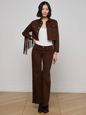A woman stands indoors wearing the Koda Fringe Jean Jacket over a white top, paired with brown high-waisted wide-leg pants and brown boots, posing against a plain light-colored wall and wood floor.
