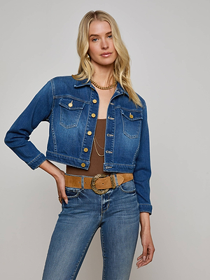 A woman with long blonde hair wears the L'AGENCE Koda Cropped Jean Jacket over a brown top, blue jeans, and gold necklaces, standing against a plain light background.