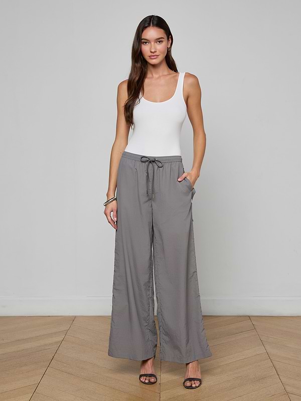 A woman stands against a white background, wearing a white tank top, the Valkyrie Snap Pant in gray featuring a wide-leg silhouette and drawstring waist, paired with black heeled sandals and her hands in her pockets.