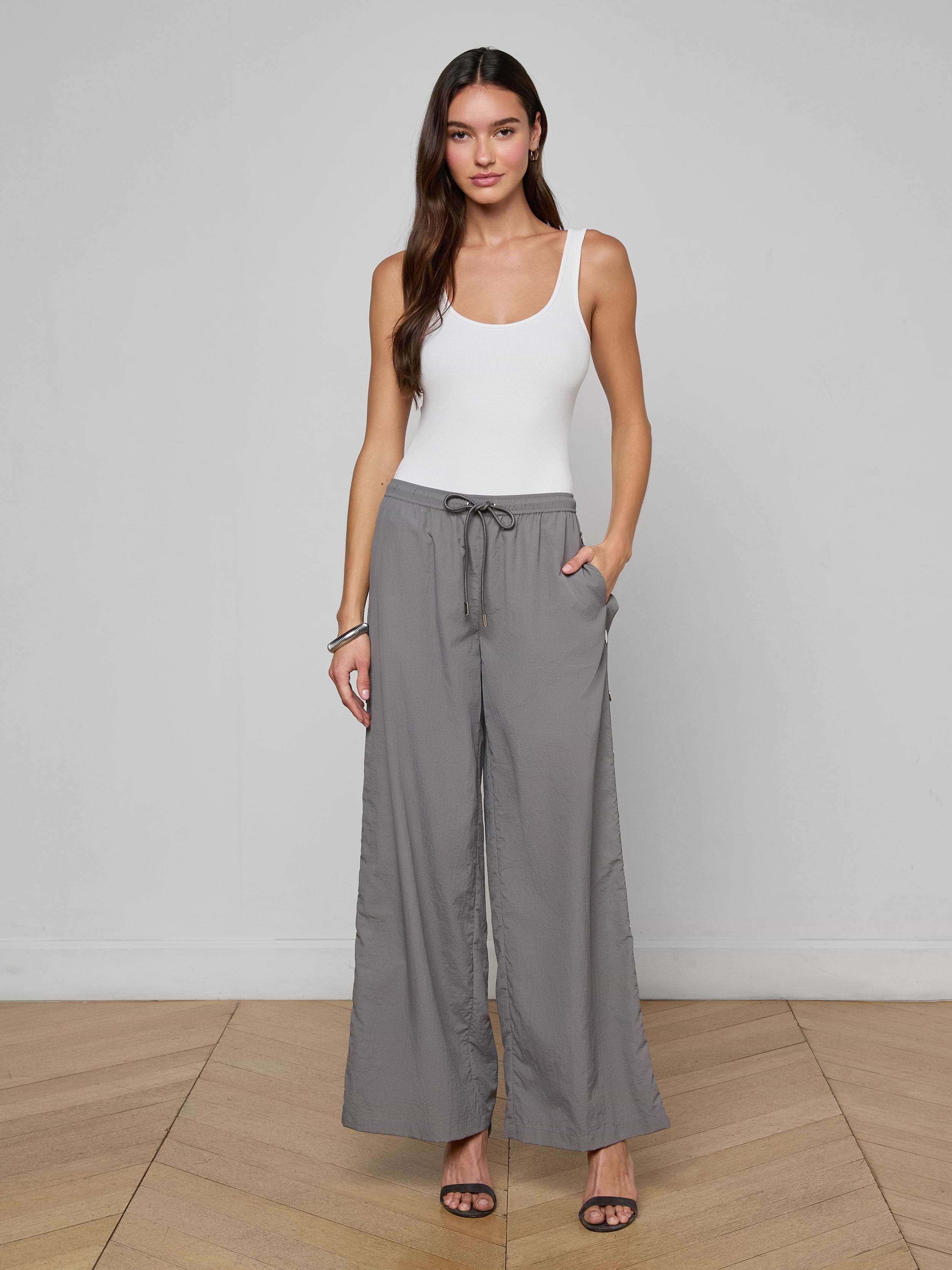 A woman stands against a white background, wearing a white tank top, the Valkyrie Snap Pant in gray featuring a wide-leg silhouette and drawstring waist, paired with black heeled sandals and her hands in her pockets.