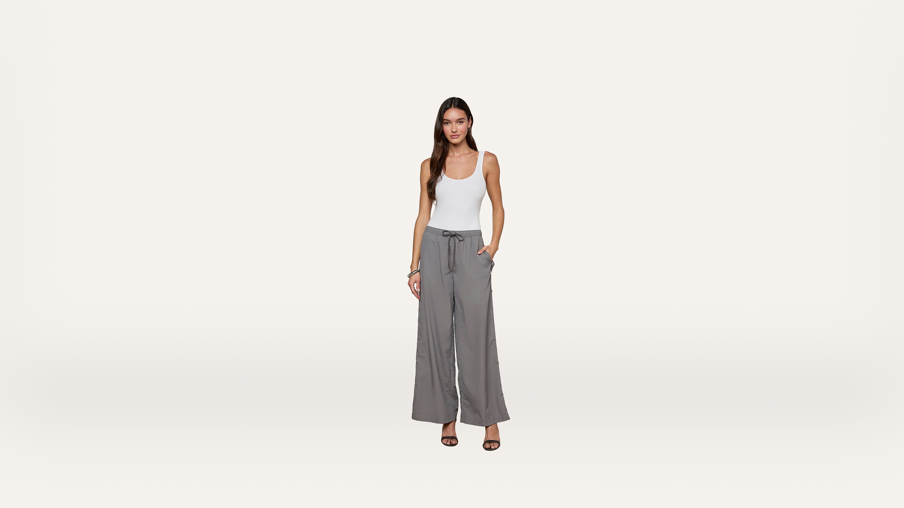 A woman stands against a plain white background, wearing a white tank top, gray wide-leg pants with a drawstring waist, and black heeled sandals. One hand is in her pocket, and she is facing forward, smiling slightly.