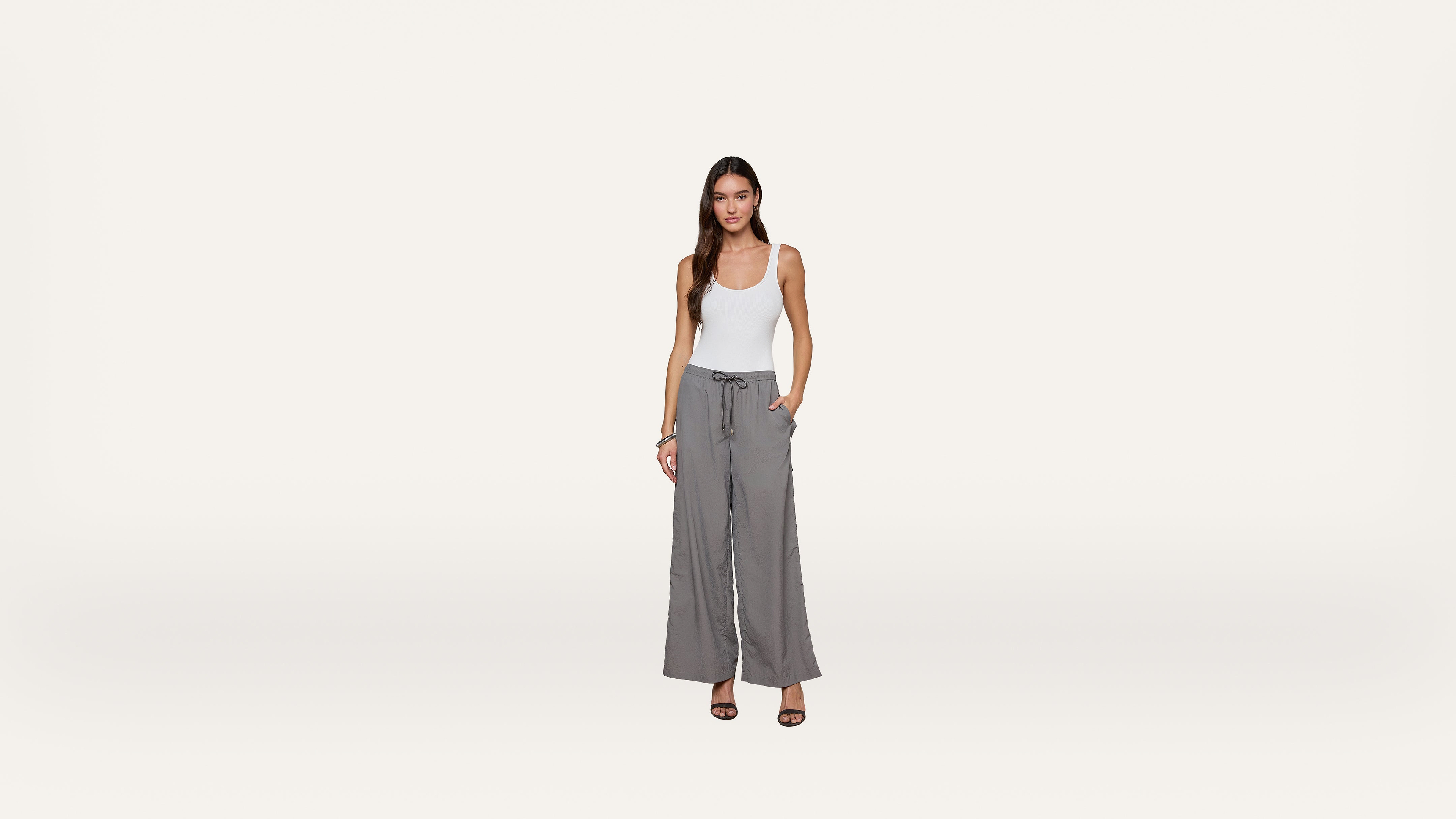 A woman stands against a plain white background, wearing a white tank top, gray wide-leg pants with a drawstring waist, and black heeled sandals. One hand is in her pocket, and she is facing forward, smiling slightly.