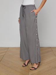 A woman wears loose grey Valkyrie Snap Pants with side buttons, paired with black high-heeled sandals and a white sleeveless top. Her left hand rests in her pocket, and the background is plain off-white.