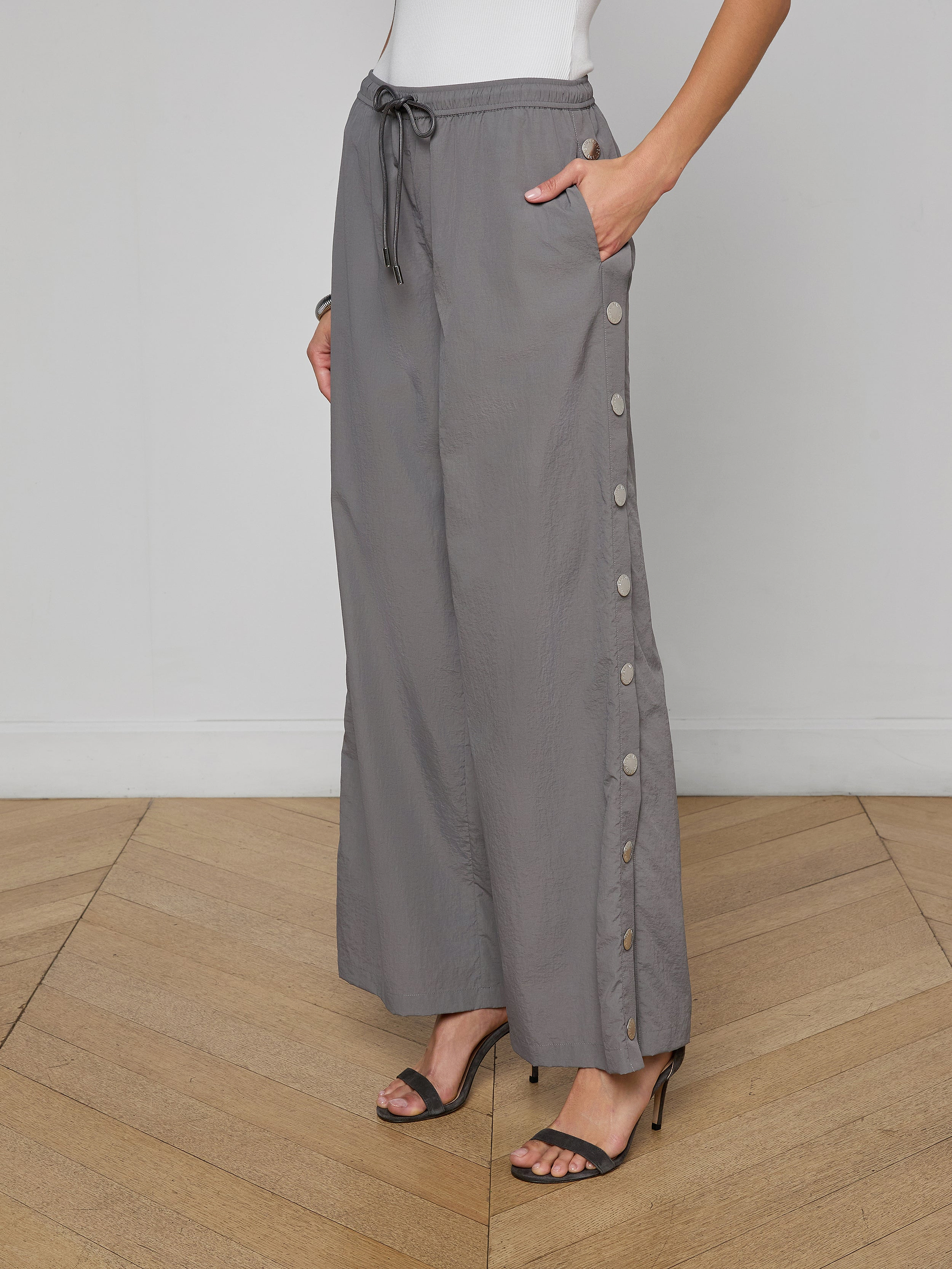 A woman wears loose grey Valkyrie Snap Pants with side buttons, paired with black high-heeled sandals and a white sleeveless top. Her left hand rests in her pocket, and the background is plain off-white.