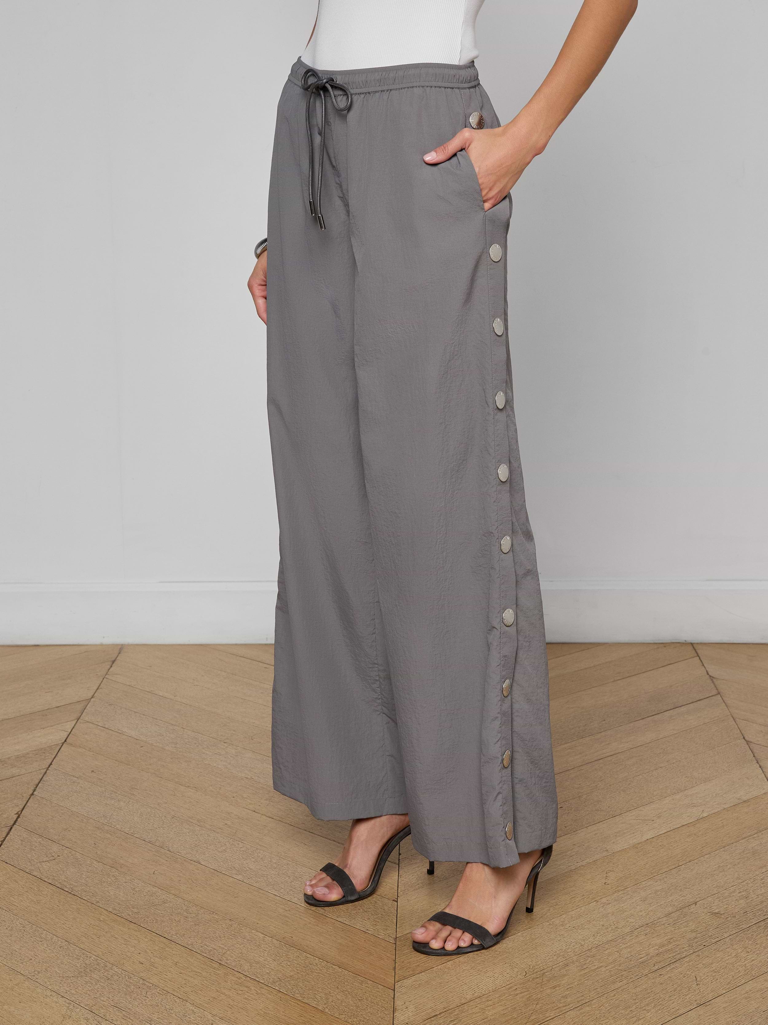 A woman wears loose grey Valkyrie Snap Pants with side buttons, paired with black high-heeled sandals and a white sleeveless top. Her left hand rests in her pocket, and the background is plain off-white.