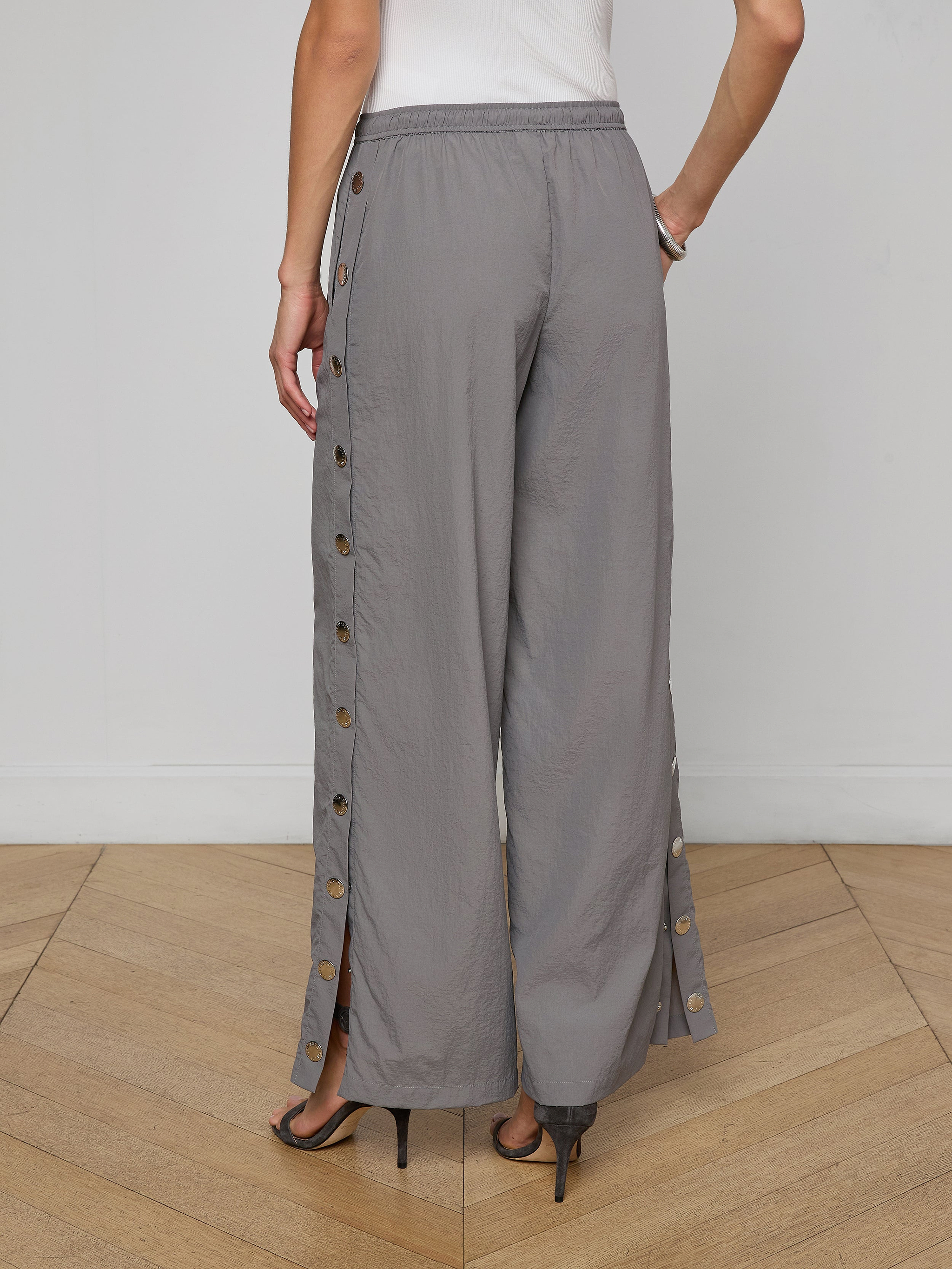 Person wearing loose gray Valkyrie Snap Pant with side buttons and black heels, standing with one hand on hip. Back view against a plain background.