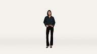 A woman stands against a plain white background wearing a dark jacket, black pants, and heeled sandals, her hands casually resting in her pockets.
