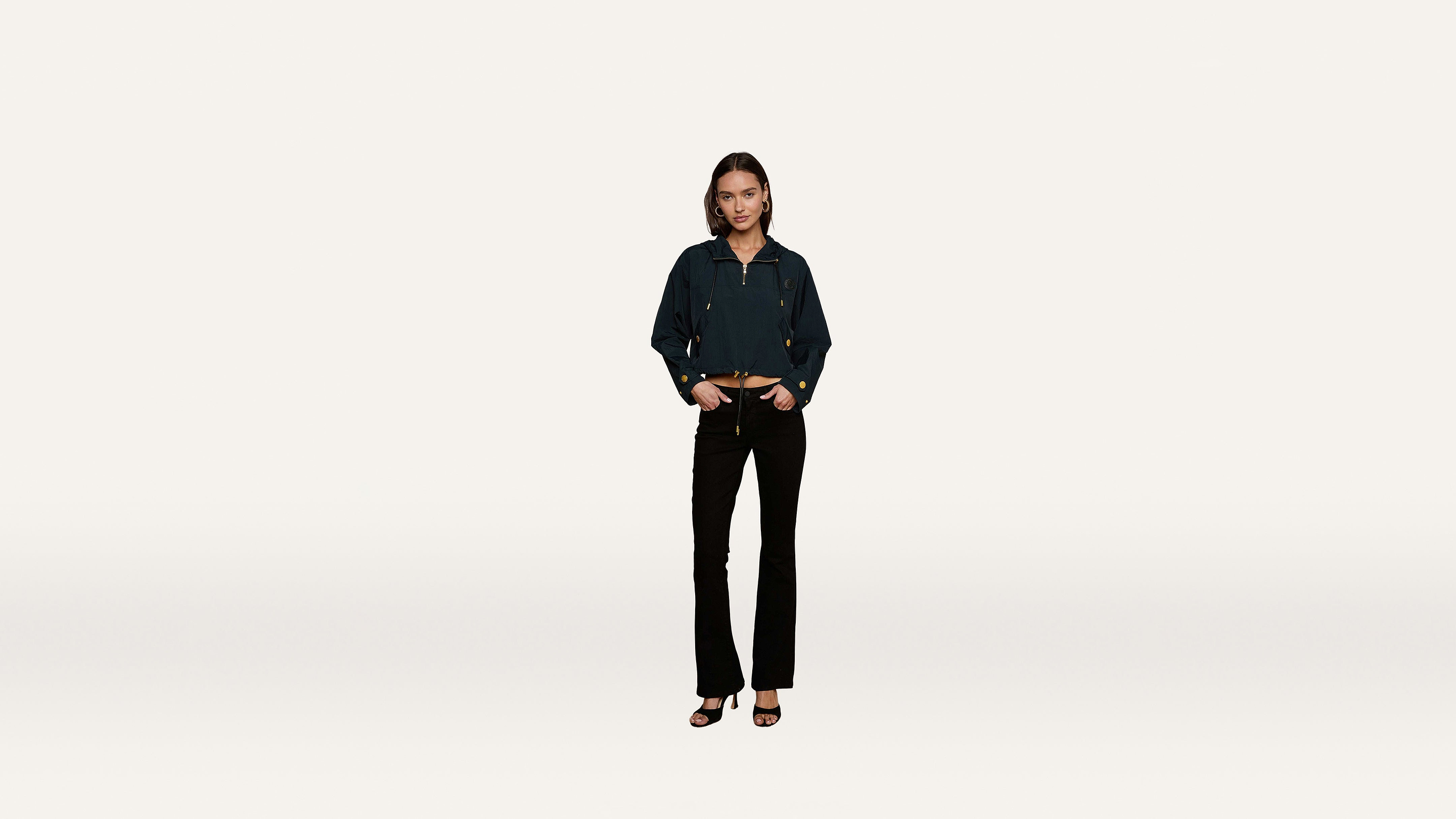 A woman stands against a plain white background wearing a dark jacket, black pants, and heeled sandals, her hands casually resting in her pockets.