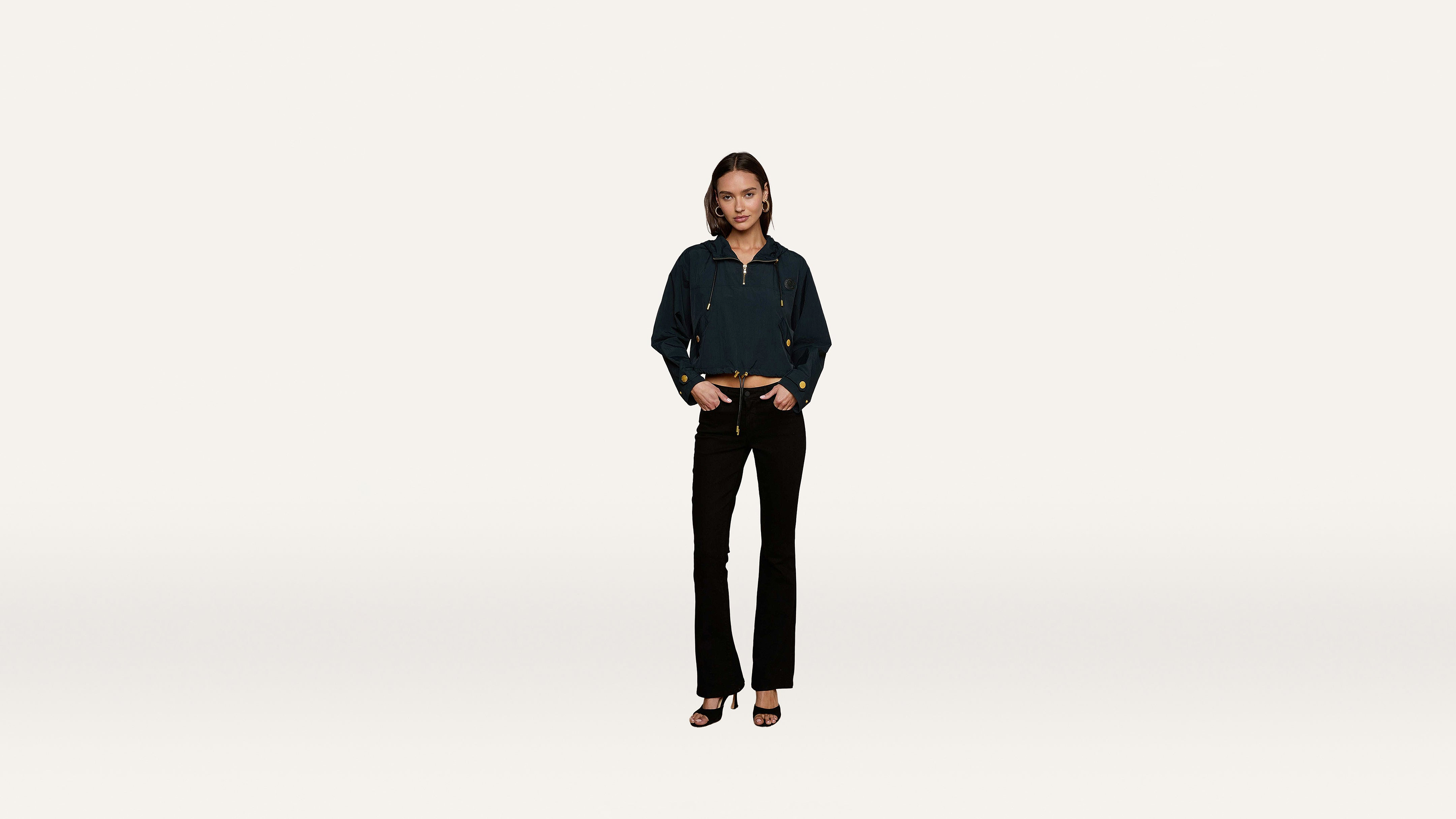 A woman stands against a plain white background wearing a dark jacket, black pants, and heeled sandals, her hands casually resting in her pockets.