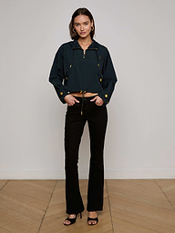 A woman with shoulder-length brown hair stands against a plain background, wearing the Bronwyn Cropped Windbreaker in dark blue, styled with black flared pants and black heeled sandals. She looks at the camera with a neutral expression.