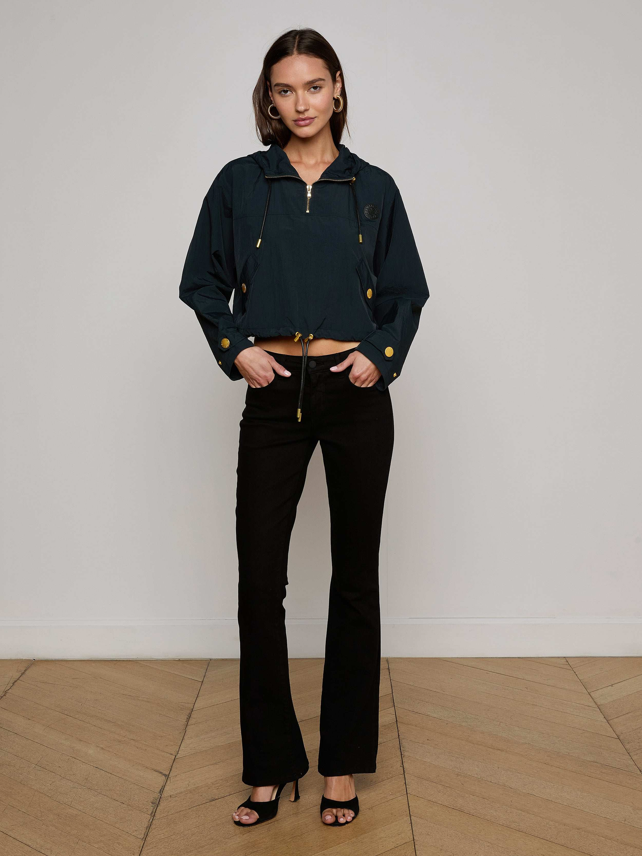 A woman with shoulder-length brown hair stands against a plain background, wearing the Bronwyn Cropped Windbreaker in dark blue, styled with black flared pants and black heeled sandals. She looks at the camera with a neutral expression.