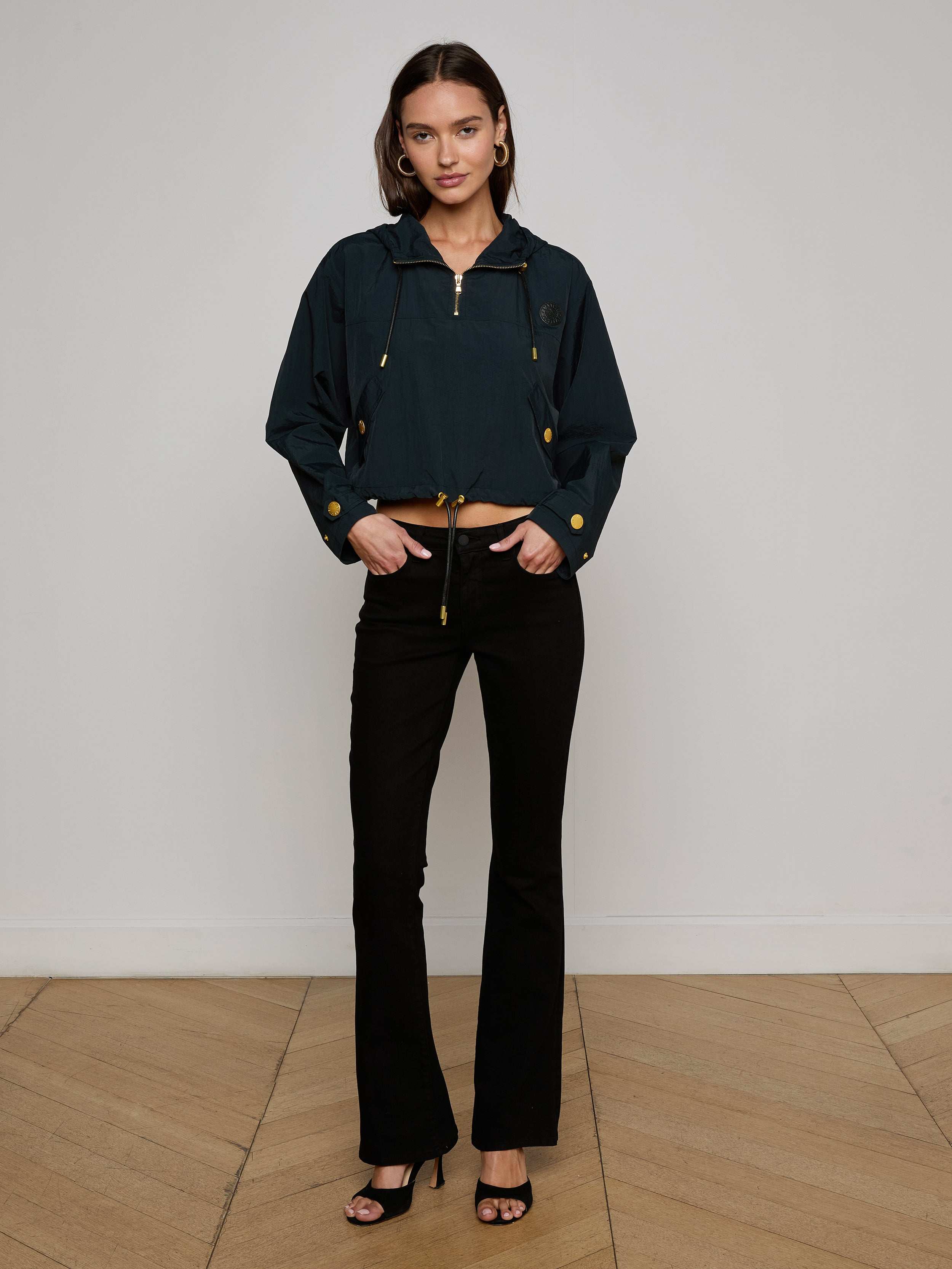 A woman with shoulder-length brown hair stands against a plain background, wearing the Bronwyn Cropped Windbreaker in dark blue, styled with black flared pants and black heeled sandals. She looks at the camera with a neutral expression.