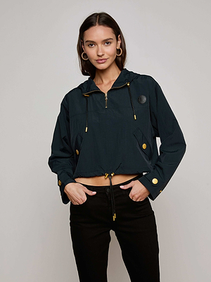 A woman in the Bronwyn Cropped Windbreaker, featuring gold buttons and zippers, pairs it with black pants and hoop earrings. She stands against a light background, smiling slightly with her hands in her pockets.
