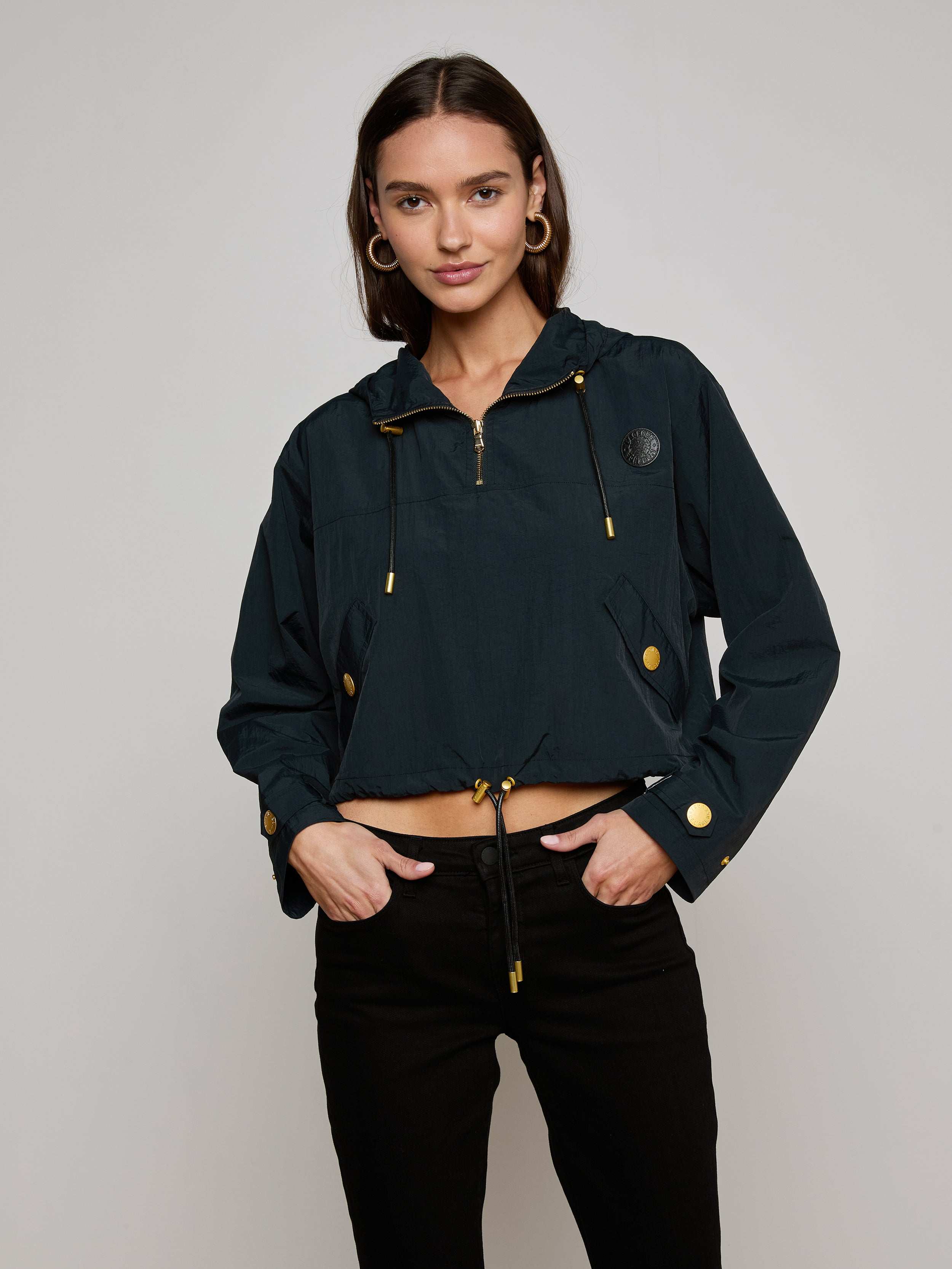 Wearing the Bronwyn Cropped Windbreaker—a dark, water-repellent jacket featuring gold buttons and zippers—a woman stands with one hand in the pocket of her black pants, smiling slightly against a plain light background.