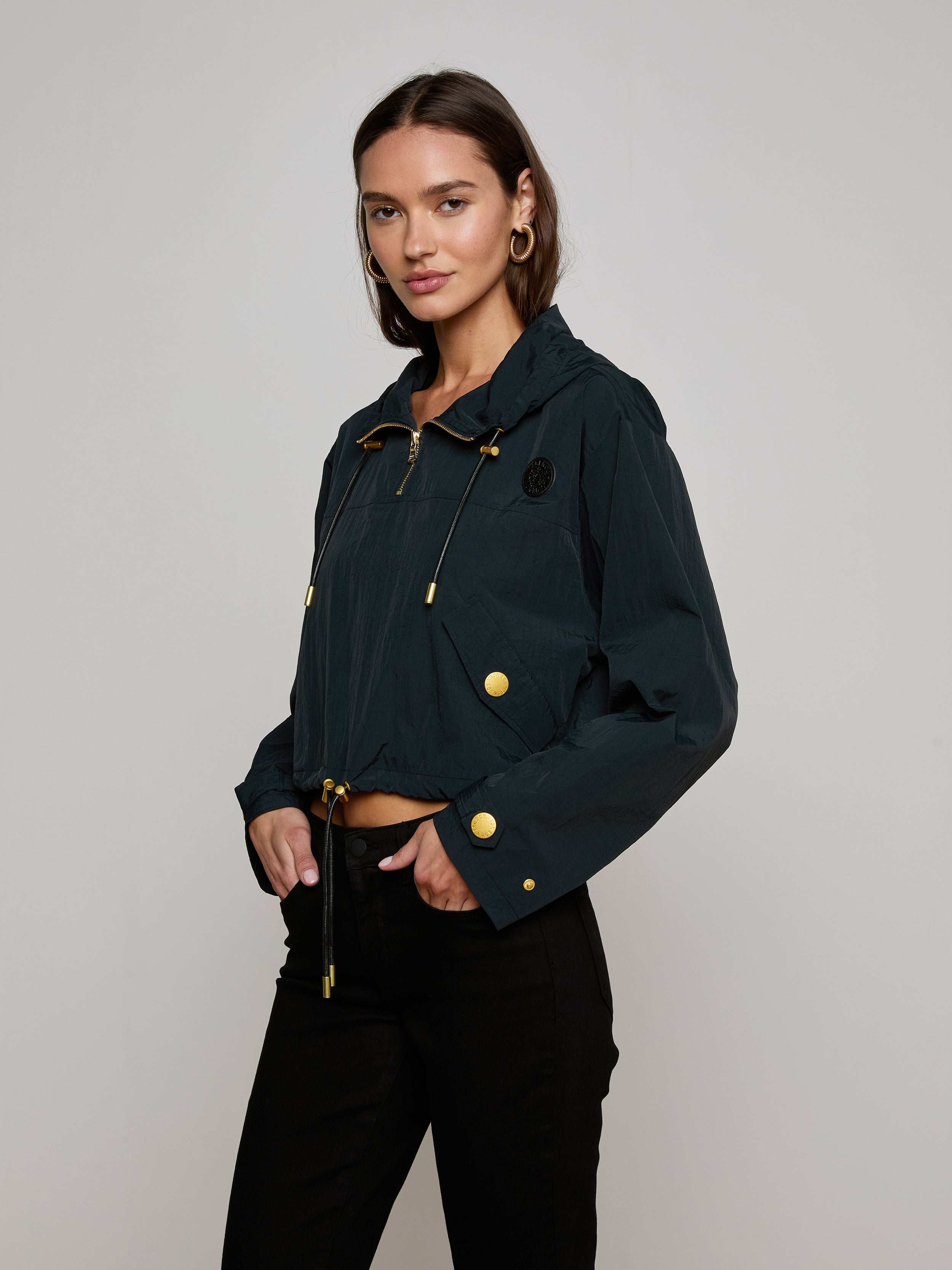 A woman wears the Bronwyn Cropped Windbreaker in dark green, paired with black pants, standing against a plain light background and looking at the camera with a neutral expression.