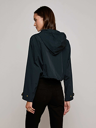 A woman in the Bronwyn Cropped Windbreaker—a dark, water-repellent hooded jacket—paired with black pants stands facing away, turning her head slightly to the side against a plain off-white background.