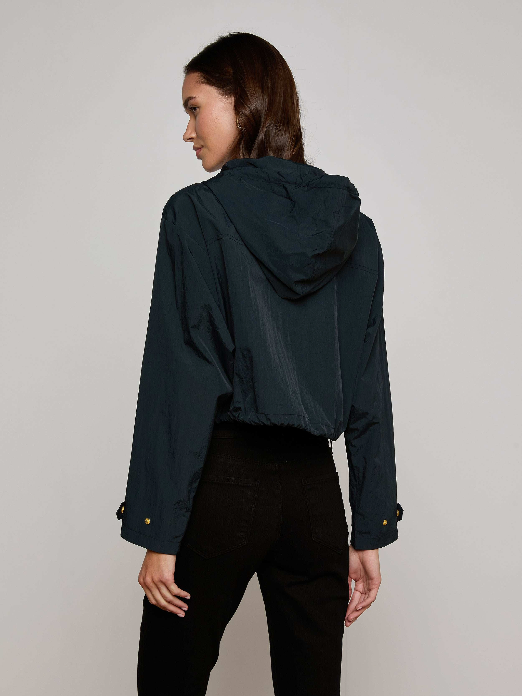 A woman in the Bronwyn Cropped Windbreaker—a dark, water-repellent hooded jacket—paired with black pants stands facing away, turning her head slightly to the side against a plain off-white background.