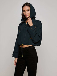 Wearing the Bronwyn Cropped Windbreaker, a dark hooded, water-repellent jacket with an elevated windbreaker design, and black pants, a woman stands against a plain light background looking at the camera with one hand holding her hood.