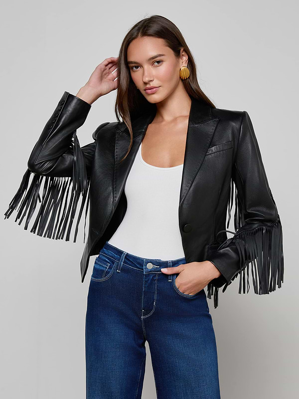 A woman wearing the Vashti Fringed Leather Blazer, styled with a white top and blue jeans, stands against a plain light background.
