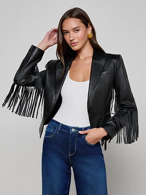 A woman models the L'AGENCE Vashti Fringed Leather Blazer in black, styled with a white top and blue jeans, posing with one hand near her head against a plain light gray background.