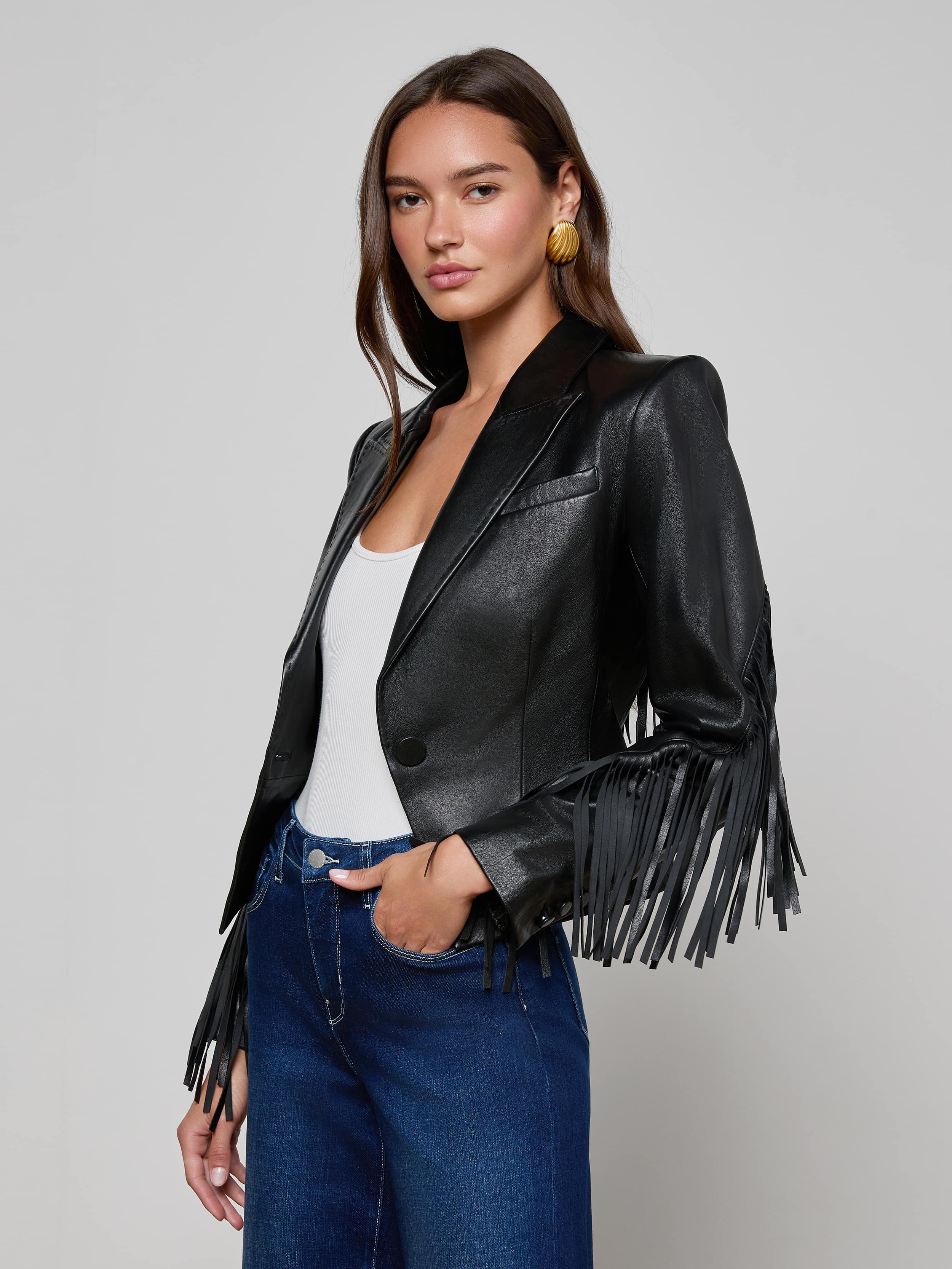 A woman wearing the Vashti Fringed Leather Blazer, crafted from Italian lambskin leather with statement fringe on the sleeves, pairs it with a white top and blue jeans as she stands against a plain light background.
