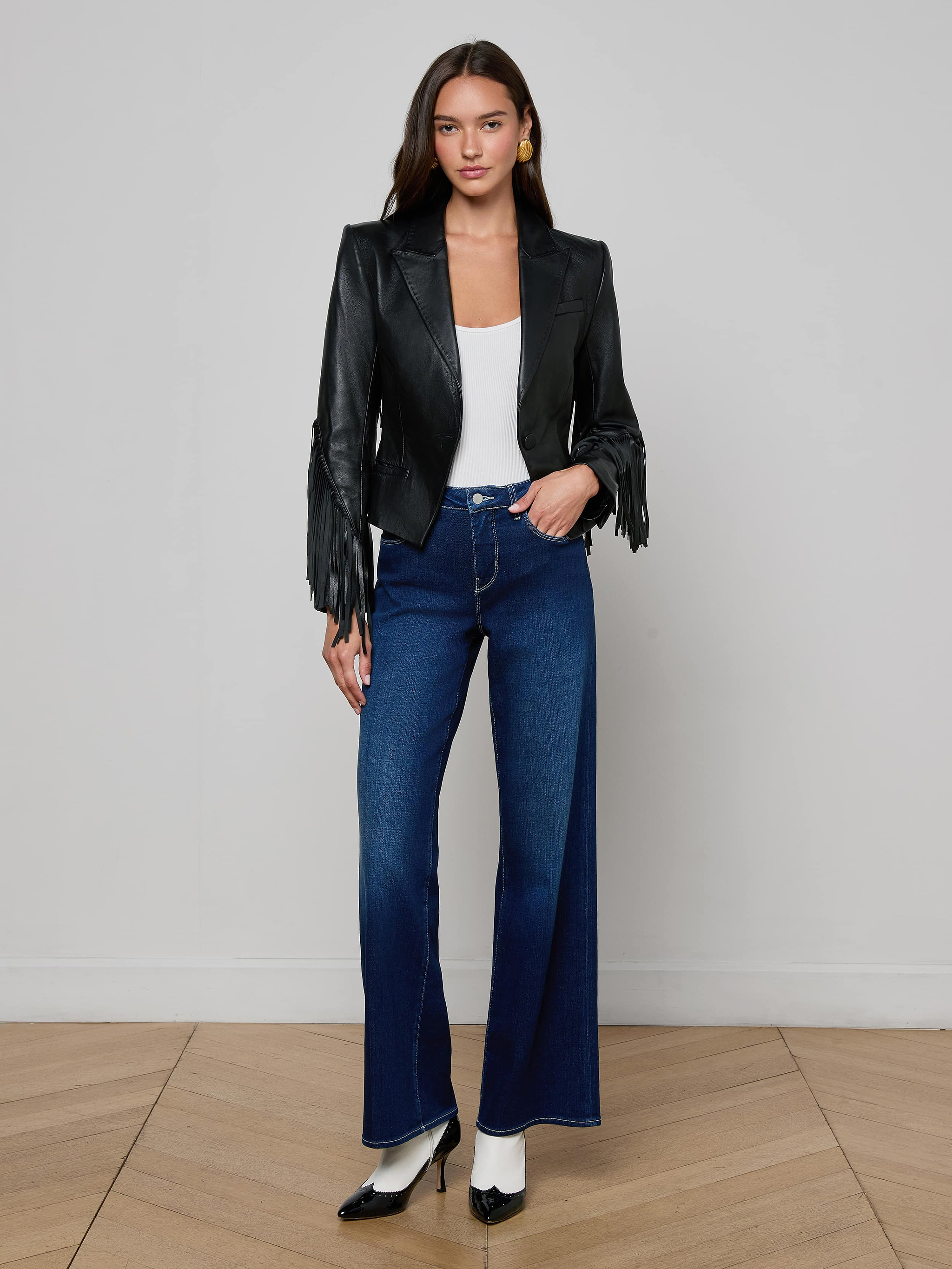 A woman stands against a plain background, wearing the Vashti Fringed Leather Blazer crafted from Italian lambskin with statement fringe, paired with a white top, high-waisted blue wide-leg jeans, and black-and-white heels.