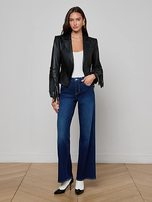 A woman stands indoors on a wooden floor, wearing the L'AGENCE Vashti Fringed Leather Blazer, a white top, high-waisted blue flared jeans, white socks, and black high-heeled shoes.