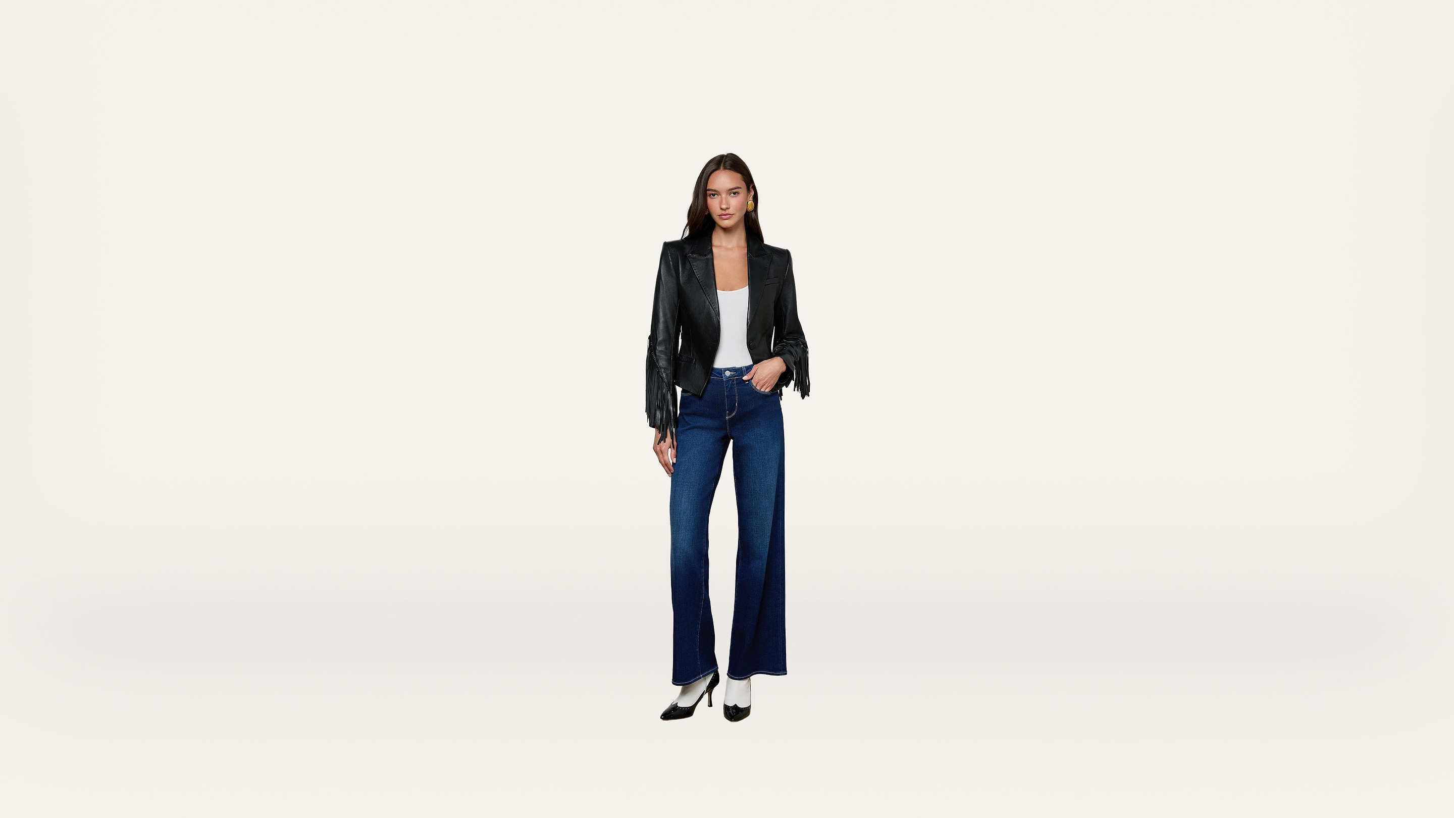 A woman stands facing forward, wearing a black jacket, white top, wide-leg blue jeans, and black heels, against a plain white background.
