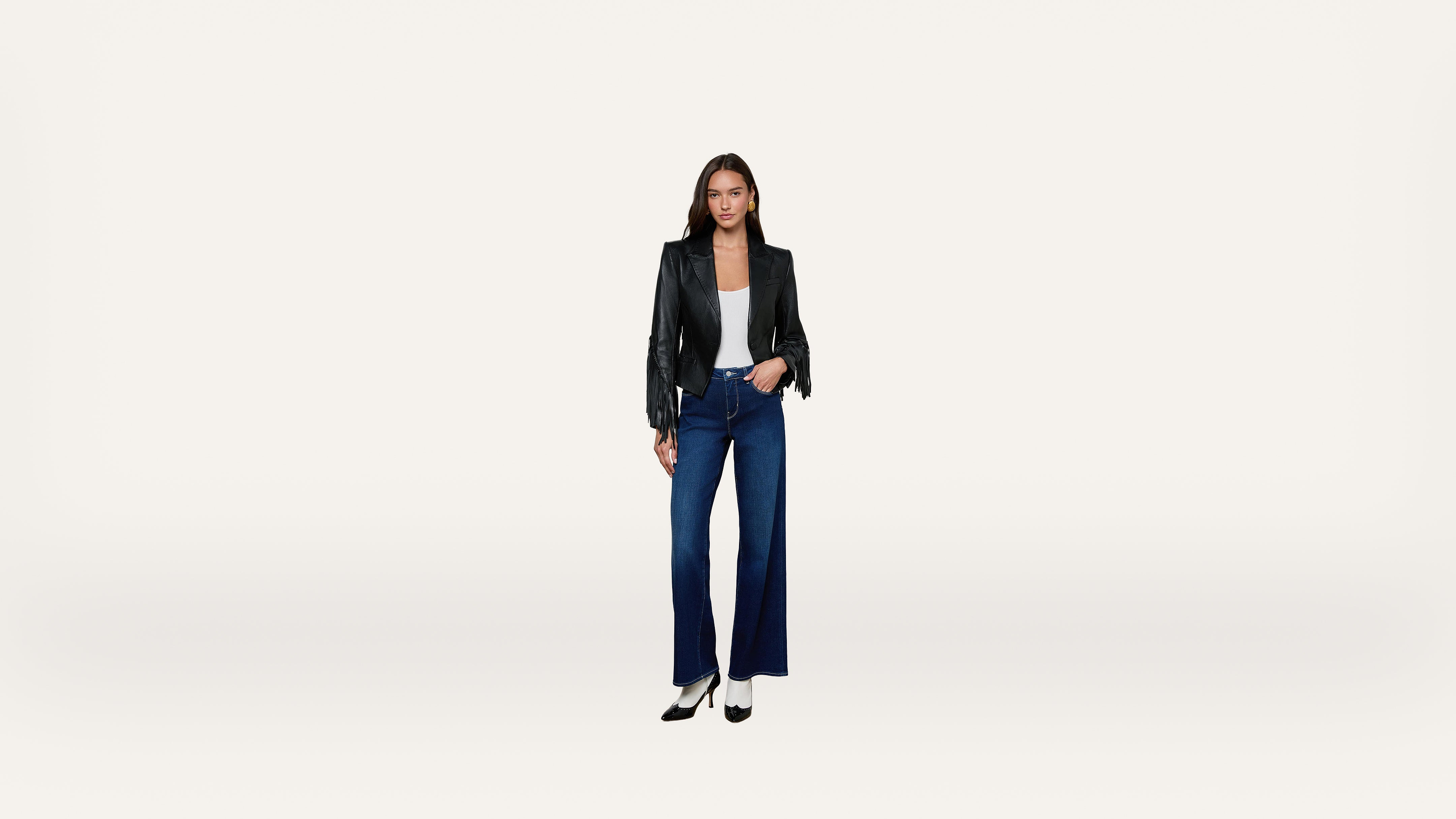 A woman stands facing forward, wearing a black jacket, white top, wide-leg blue jeans, and black heels, against a plain white background.