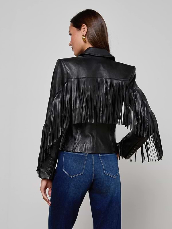 A woman stands facing away against a plain, light background, wearing the Vashti Fringed Leather Blazer—a black Italian lambskin leather jacket featuring statement fringe detailing on the back and arms—paired with blue jeans.
