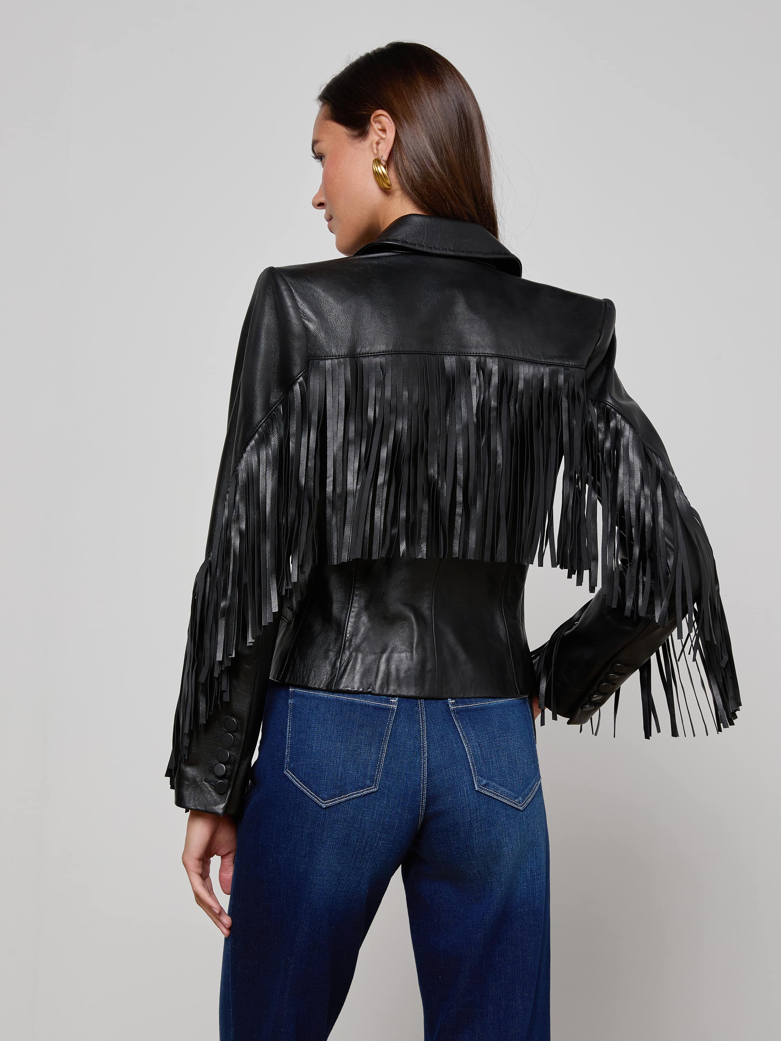 A woman stands facing away against a plain, light background, wearing the Vashti Fringed Leather Blazer—a black Italian lambskin leather jacket featuring statement fringe detailing on the back and arms—paired with blue jeans.
