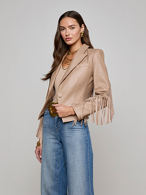 A woman with long brown hair wears the Vashti Fringed Leather Blazer in beige Italian lambskin over a matching top, paired with high-waisted blue jeans, gold jewelry, and a brown belt, showcasing her hourglass figure against a plain light background.