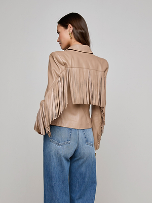 A woman with an hourglass figure, facing away, wears the Vashti Fringed Leather Blazer in tan Italian lambskin with long fringe on the back and sleeves, paired with blue jeans. The background is plain and light gray.