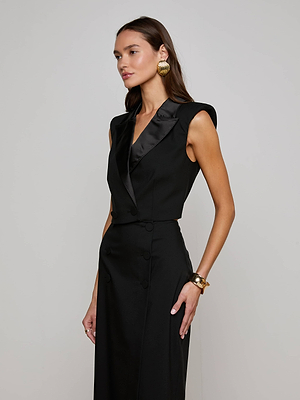 A woman stands against a plain background wearing the Hollis Vest by L'AGENCE, a sleeveless black dress with satin lapels, styled with gold hoop earrings and a gold bracelet, looking to the side with her hair down.