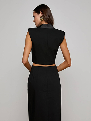 A woman with long brown hair is seen from the back, wearing the L'AGENCE Hollis Vest and a matching high-waisted black skirt, finished with gold statement earrings.